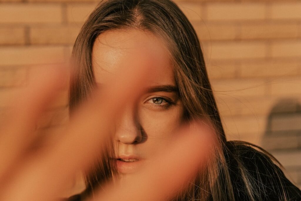 A person with dark brown hair peers around their hand.